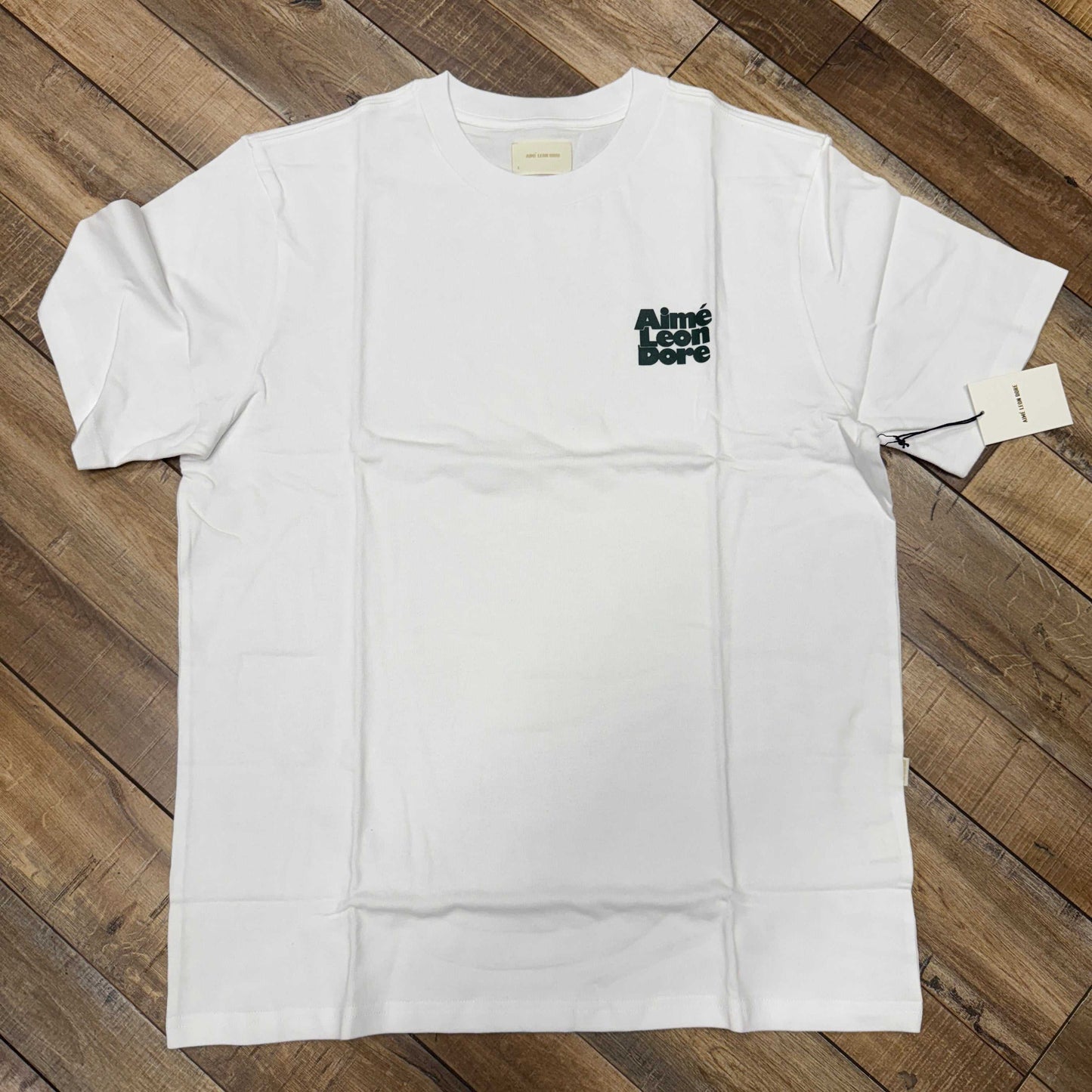 Aimé Leon Dore Stacked Logo Tee Bright White on wooden floor.