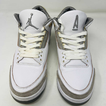 The Holy Ground Jordan 3 Retro SP A Ma Maniére (Womens) sneakers in white and gray, priced at $450, are displayed atop their box. They feature textured leather, suede accents, and include a pack of cream-colored extra laces for customization.
