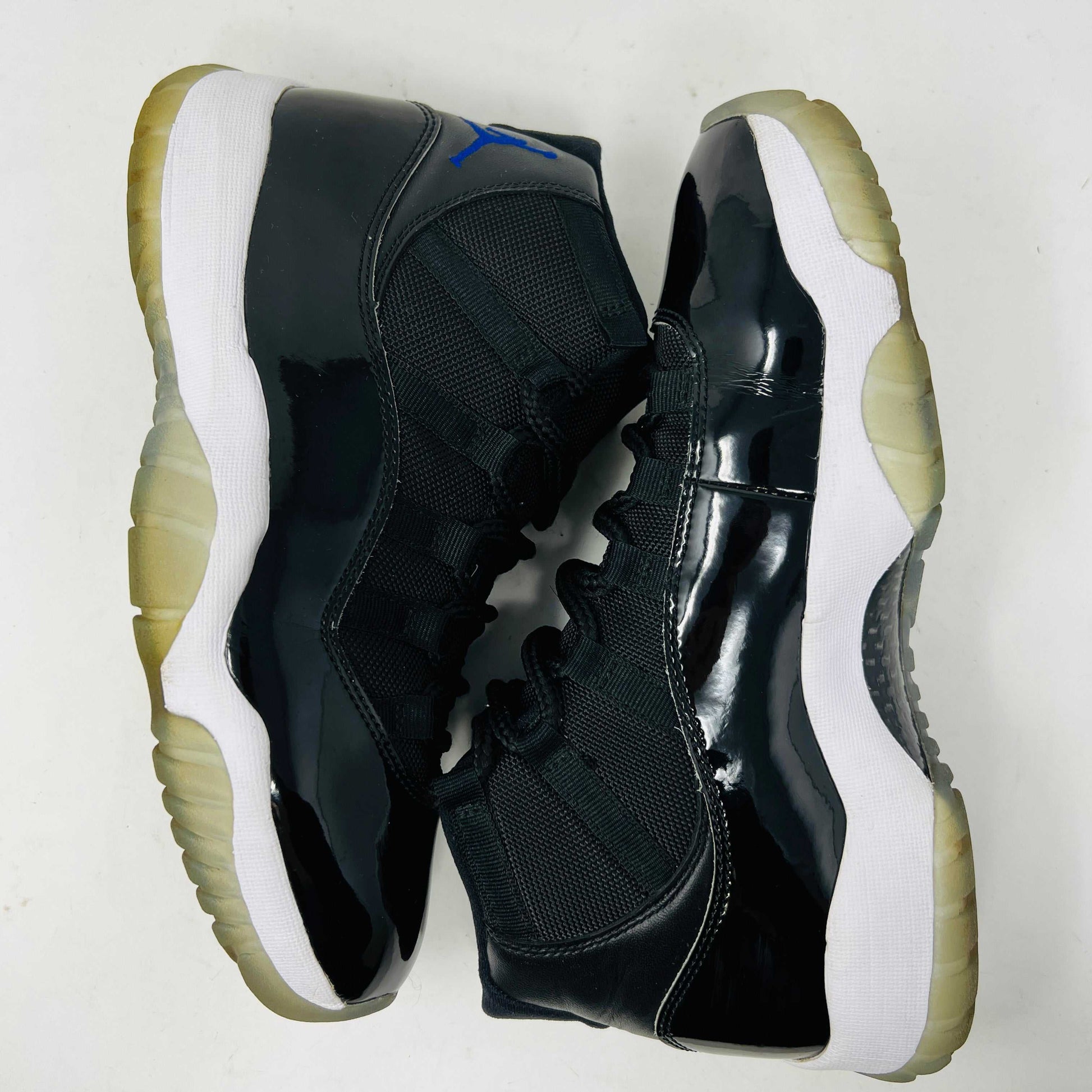 A Holy Ground Jordan 11 Retro Space Jam (2016) sneaker, featuring a glossy black patent leather upper with a blue Jumpman logo, sits on its original box. It has a white midsole and translucent outsole and is guaranteed 100% authentic with detailed sizing information on the box.