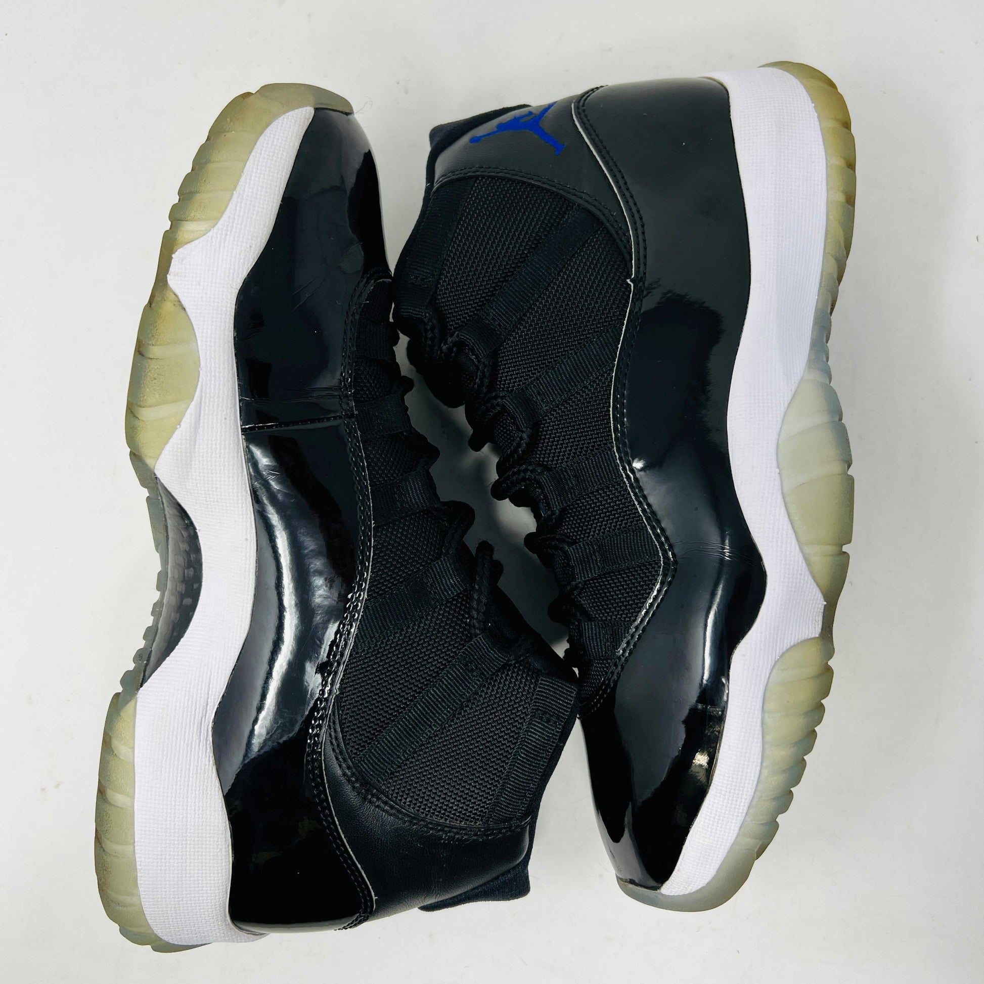 A Holy Ground Jordan 11 Retro Space Jam (2016) sneaker, featuring a glossy black patent leather upper with a blue Jumpman logo, sits on its original box. It has a white midsole and translucent outsole and is guaranteed 100% authentic with detailed sizing information on the box.