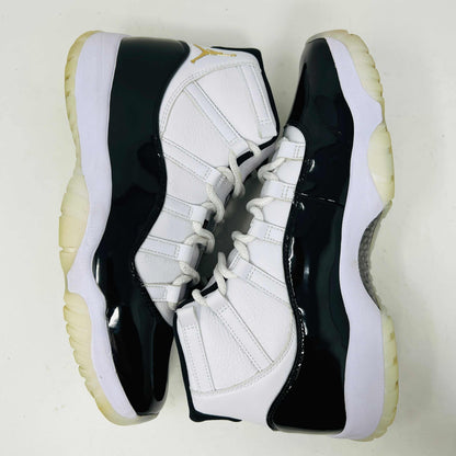 The Jordan 11 Retro DMP Gratitude (2023) sneaker, featuring white mesh, black patent leather, a translucent sole, and a gold Jumpman logo on the heel, rests elegantly on its box labeled with size and model details.