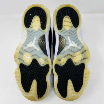 The Jordan 11 Retro DMP Gratitude (2023) sneaker, featuring white mesh, black patent leather, a translucent sole, and a gold Jumpman logo on the heel, rests elegantly on its box labeled with size and model details.