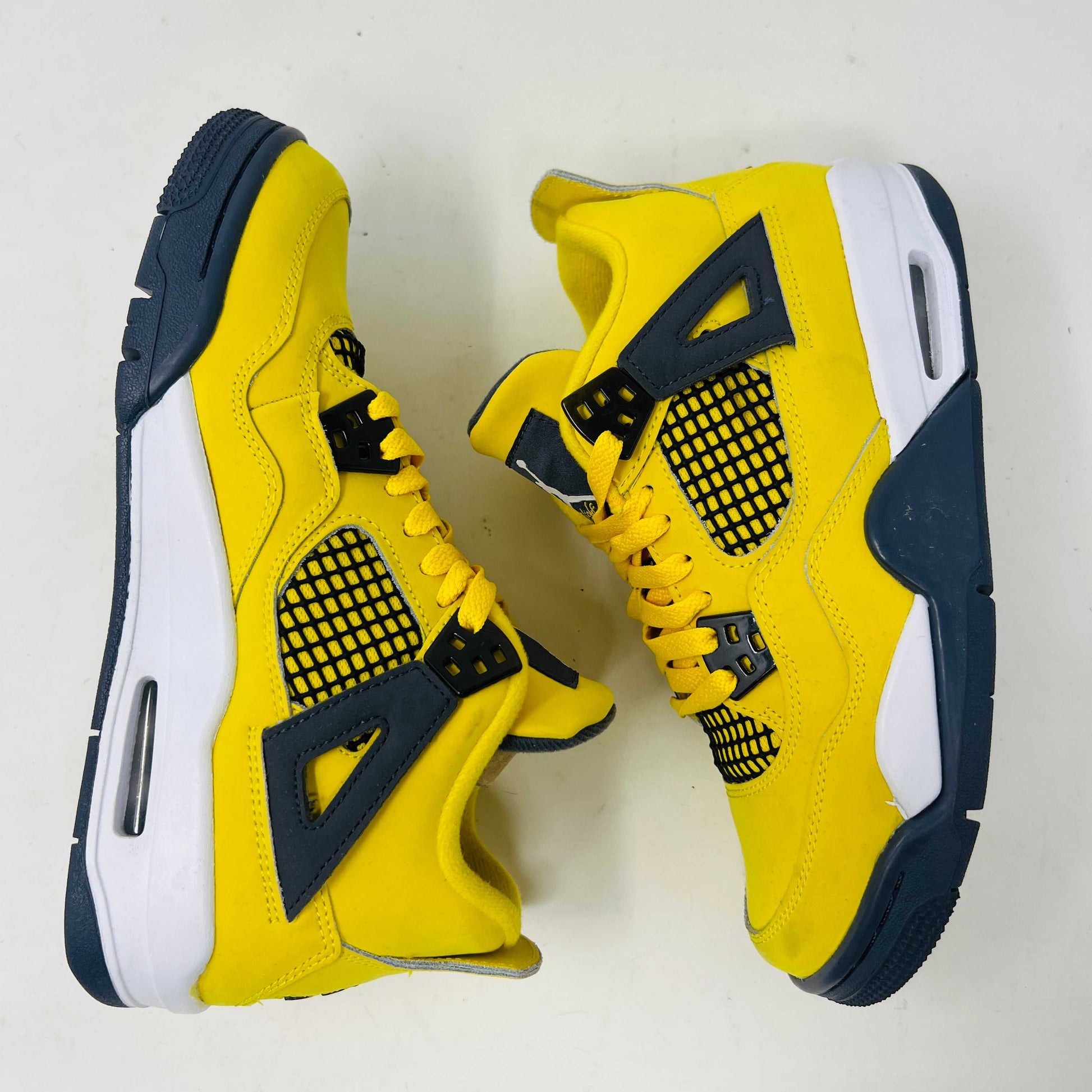 A genuine pair of Jordan 4 Retro Lightning (2021) (GS) in yellow and navy sits on a matching box with a speckled gray base, featuring mesh details, white accents, and the iconic Jumpman logo alongside product info.