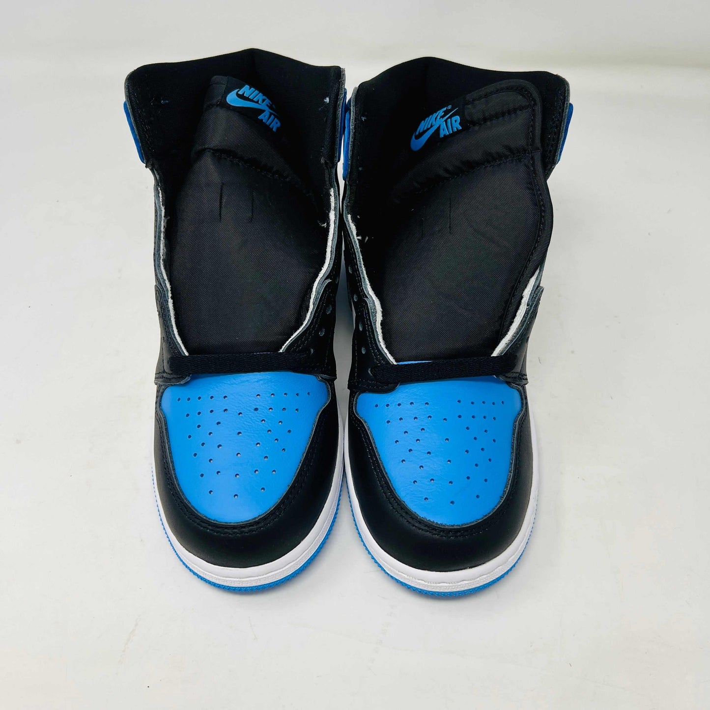 A pair of 100% authentic Jordan 1 Retro High OG UNC Toe (GS) sneakers in black, white, and blue sits on a matching blue box. The shoes feature the iconic black Nike swoosh logo on the side.