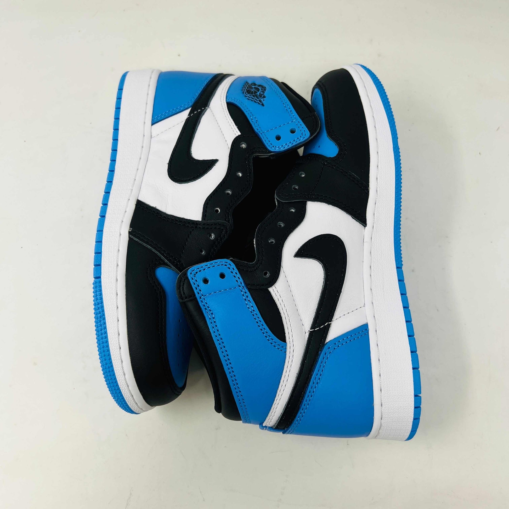 A pair of 100% authentic Jordan 1 Retro High OG UNC Toe (GS) sneakers in black, white, and blue sits on a matching blue box. The shoes feature the iconic black Nike swoosh logo on the side.