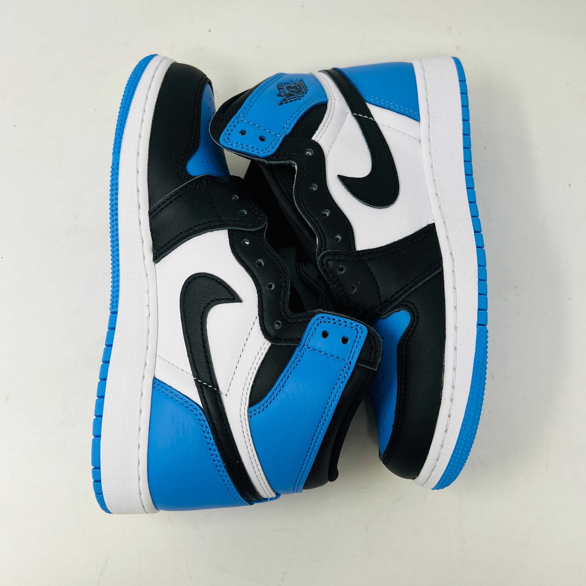 A pair of 100% authentic Jordan 1 Retro High OG UNC Toe (GS) sneakers in black, white, and blue sits on a matching blue box. The shoes feature the iconic black Nike swoosh logo on the side.