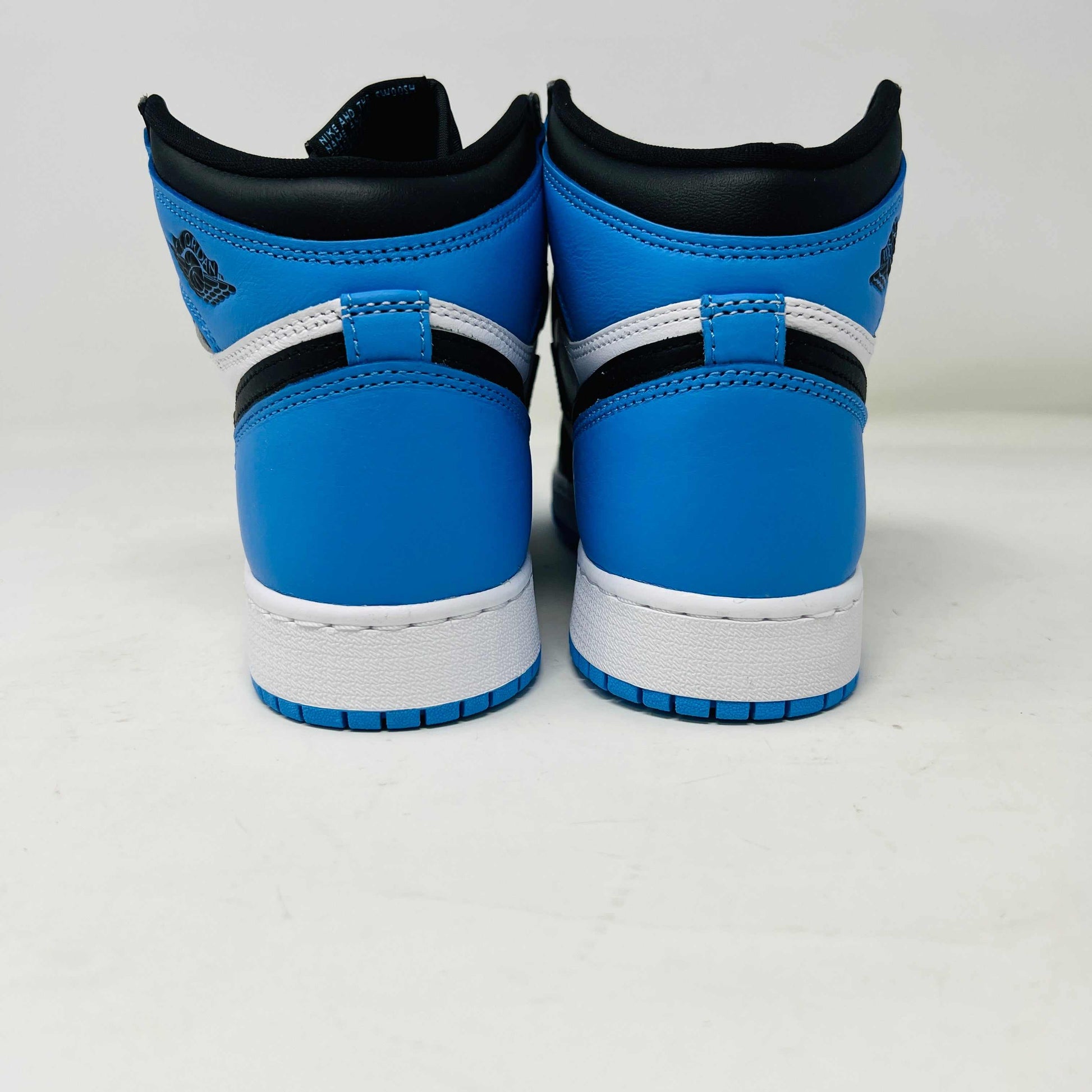 A pair of 100% authentic Jordan 1 Retro High OG UNC Toe (GS) sneakers in black, white, and blue sits on a matching blue box. The shoes feature the iconic black Nike swoosh logo on the side.