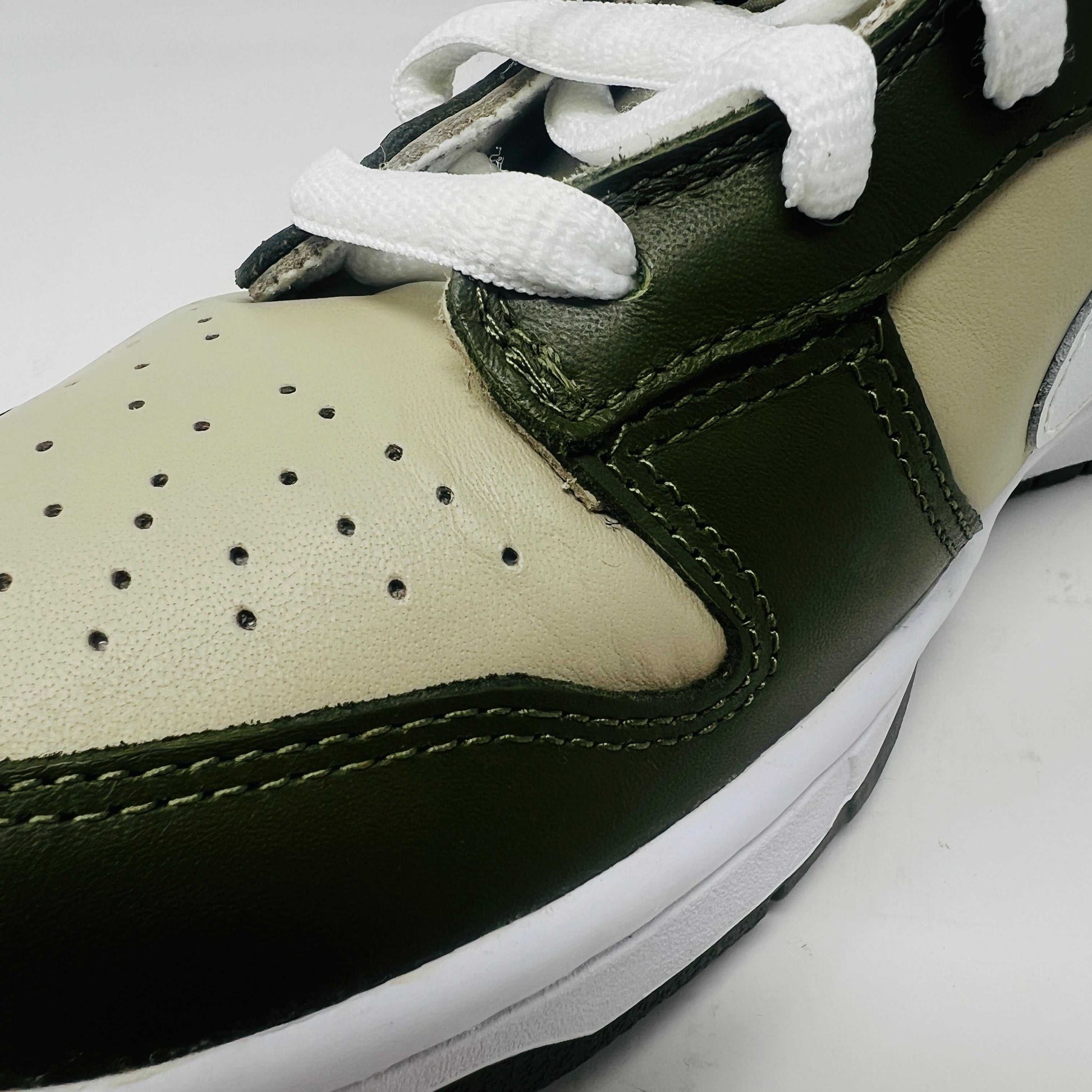 A Nike Dunk Low Light Stone shoe in green and beige with brand new white laces is displayed atop an orange Nike box. Extra white laces are in front, ensuring this 100% authentic pair from the Nike Dunk collection is ready to step out in style.