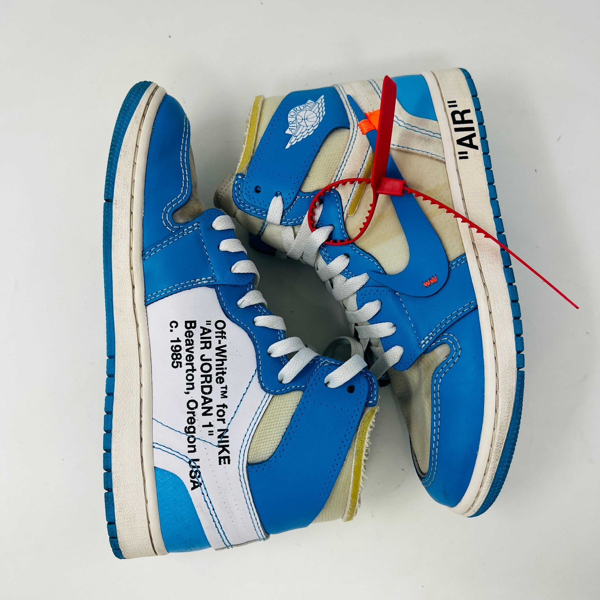 The Jordan 1 Retro High Off-White University Blue, 100% authentic, features clean blue and white uppers with AIR on the sole, orange and blue laces, and a signature red tag. Its styled atop a black and red Nike shoebox.