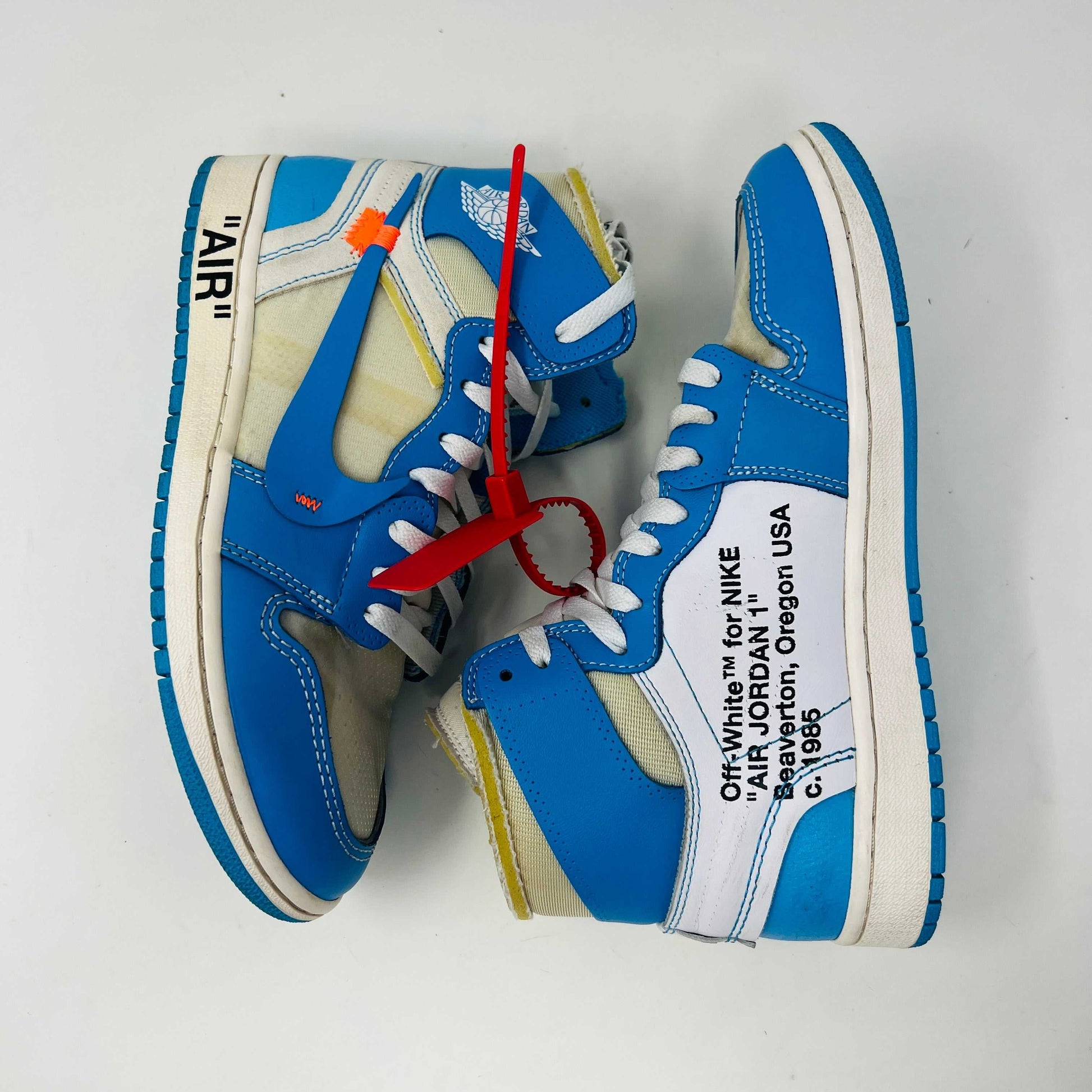 The Jordan 1 Retro High Off-White University Blue, 100% authentic, features clean blue and white uppers with AIR on the sole, orange and blue laces, and a signature red tag. Its styled atop a black and red Nike shoebox.