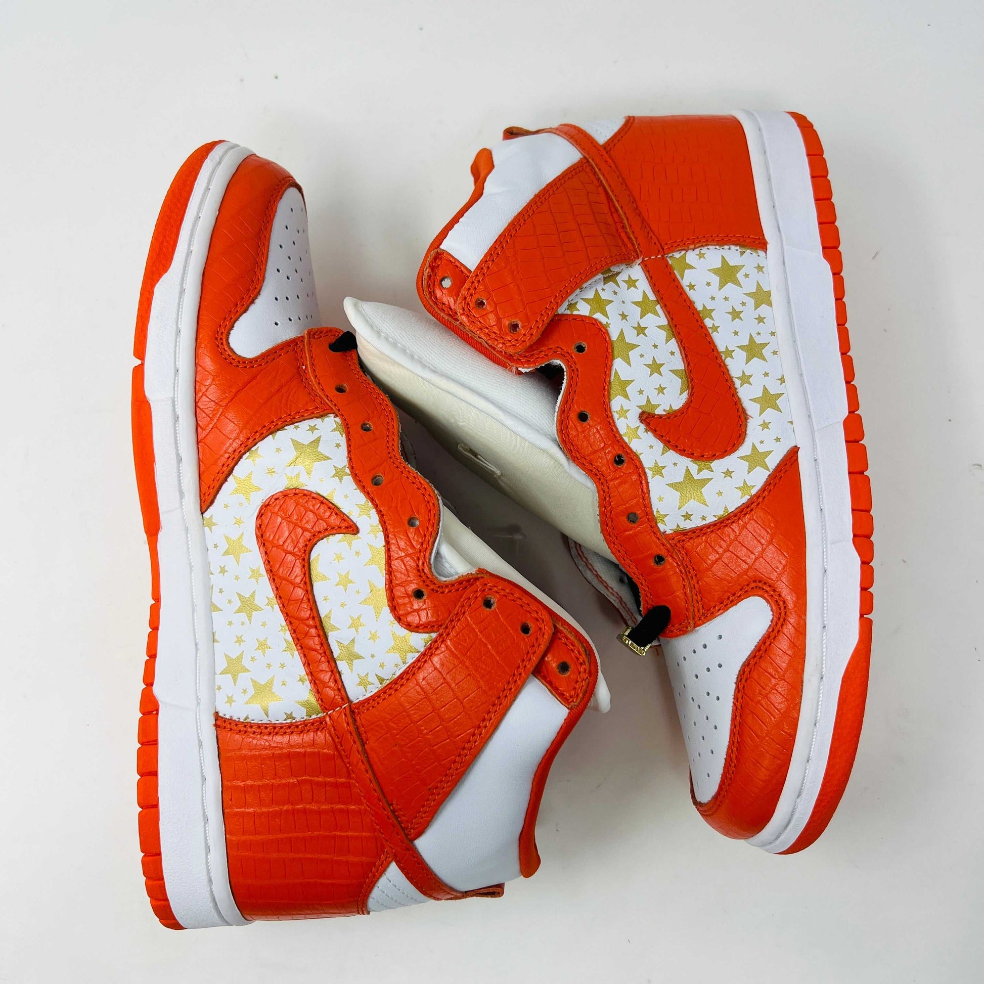 The Nike Dunk High Pro SB Supreme Orange Stars sneaker features an orange and white color scheme with star patterns on the side, a textured finish, and comes with authentic orange and white laces in a separate package.