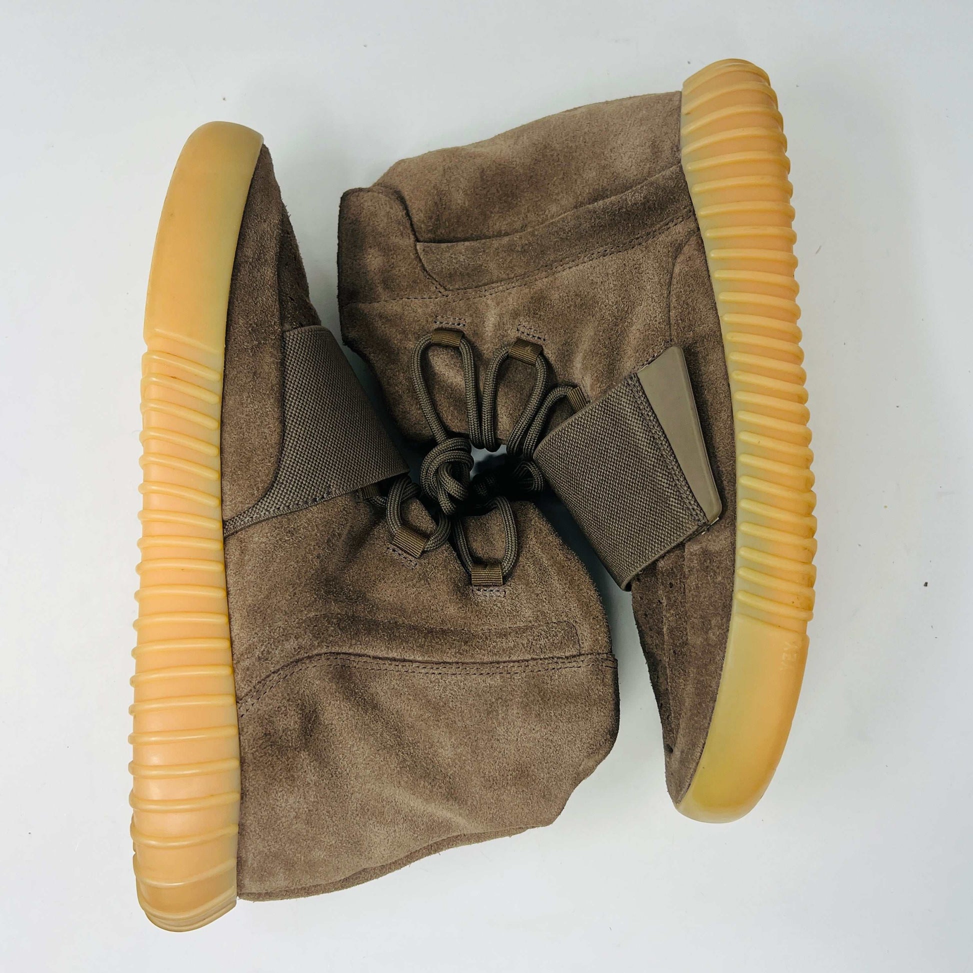 The adidas Yeezy Boost 750 Chocolate sneakers, designed by Yeezy, feature brown suede high-tops with clean uppers and thick ribbed soles. A wide elastic strap and tied laces enhance the sleek look, set against a plain white backdrop. Available on final sale.
