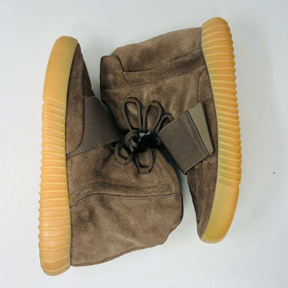 The adidas Yeezy Boost 750 Chocolate sneakers, designed by Yeezy, feature brown suede high-tops with clean uppers and thick ribbed soles. A wide elastic strap and tied laces enhance the sleek look, set against a plain white backdrop. Available on final sale.