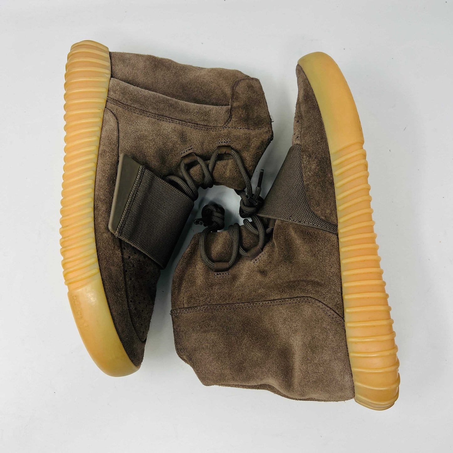 The adidas Yeezy Boost 750 Chocolate sneakers, designed by Yeezy, feature brown suede high-tops with clean uppers and thick ribbed soles. A wide elastic strap and tied laces enhance the sleek look, set against a plain white backdrop. Available on final sale.