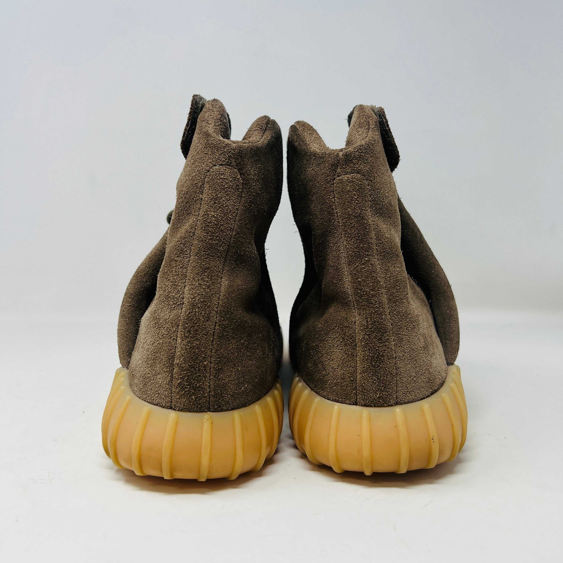 The adidas Yeezy Boost 750 Chocolate sneakers, designed by Yeezy, feature brown suede high-tops with clean uppers and thick ribbed soles. A wide elastic strap and tied laces enhance the sleek look, set against a plain white backdrop. Available on final sale.
