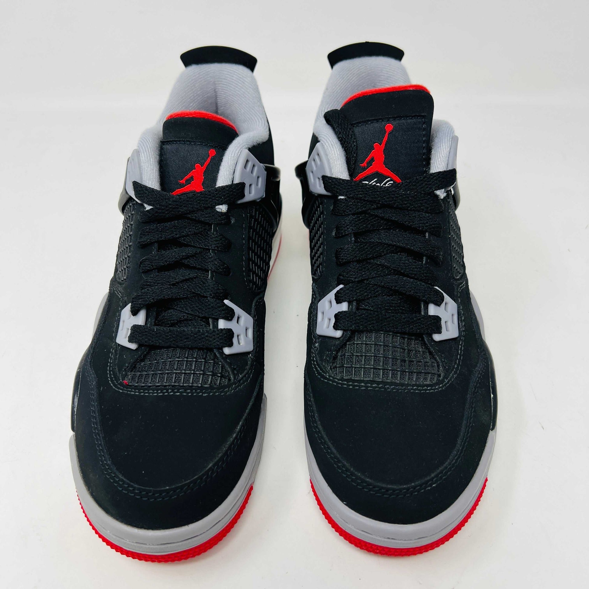 The Jordan 4 Bred 2019 GS, a black and gray sneaker with red accents, rests on a branded black box featuring the gold Jumpman logo. It boasts a visible air unit in its sole, mesh side detailing, and comes with an exclusive hang tag.
