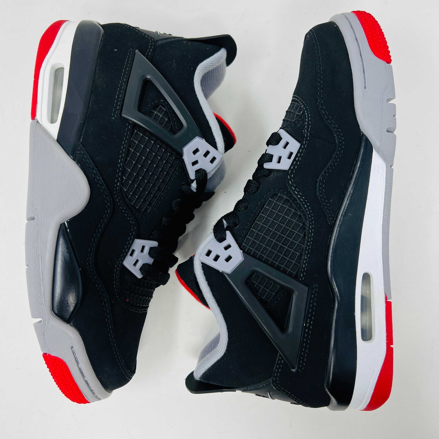 The Jordan 4 Bred 2019 GS, a black and gray sneaker with red accents, rests on a branded black box featuring the gold Jumpman logo. It boasts a visible air unit in its sole, mesh side detailing, and comes with an exclusive hang tag.