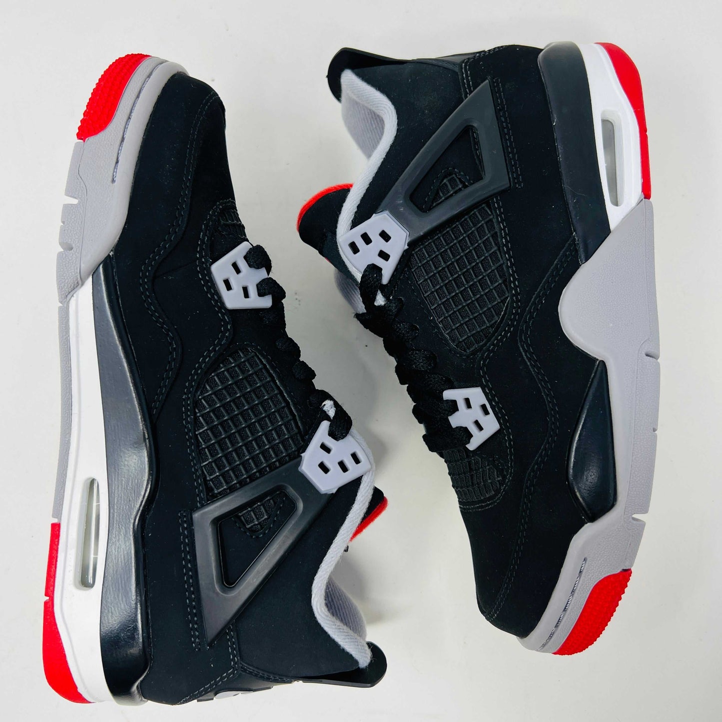 The Jordan 4 Bred 2019 GS, a black and gray sneaker with red accents, rests on a branded black box featuring the gold Jumpman logo. It boasts a visible air unit in its sole, mesh side detailing, and comes with an exclusive hang tag.