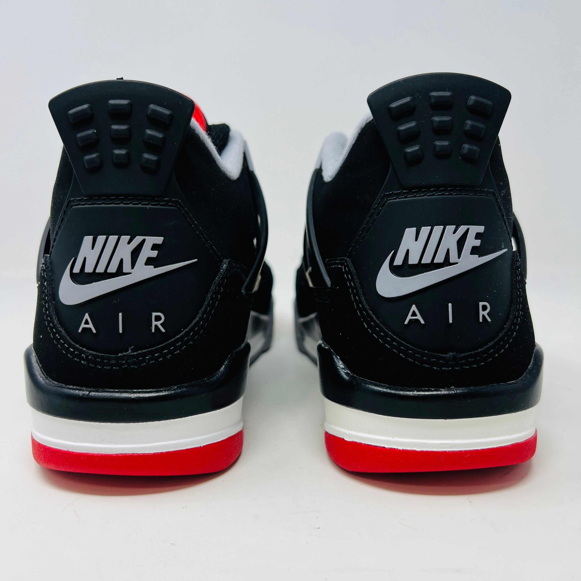 The Jordan 4 Bred 2019 GS, a black and gray sneaker with red accents, rests on a branded black box featuring the gold Jumpman logo. It boasts a visible air unit in its sole, mesh side detailing, and comes with an exclusive hang tag.