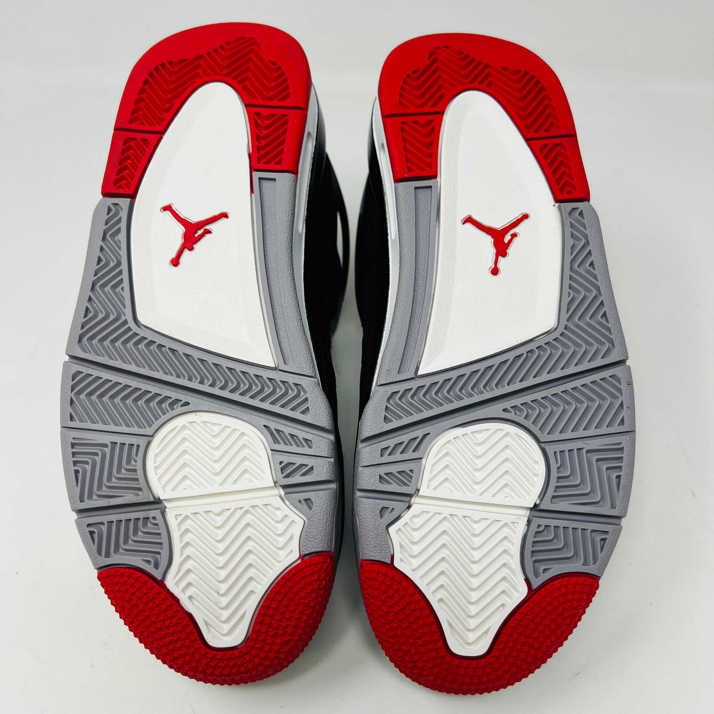 The Jordan 4 Bred 2019 GS, a black and gray sneaker with red accents, rests on a branded black box featuring the gold Jumpman logo. It boasts a visible air unit in its sole, mesh side detailing, and comes with an exclusive hang tag.
