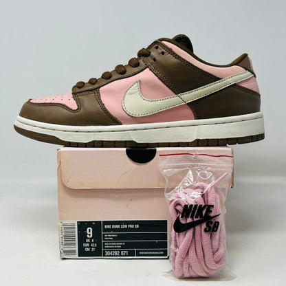 A 2005 Nike SB Dunk Low Stussy Cherry in brown and pink rests on its box, featuring a cream swoosh. The authentic shoe includes extra pink laces in a clear NIKE SB bag. The box label shows size 9.