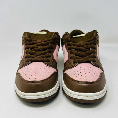 A 2005 Nike SB Dunk Low Stussy Cherry in brown and pink rests on its box, featuring a cream swoosh. The authentic shoe includes extra pink laces in a clear NIKE SB bag. The box label shows size 9.