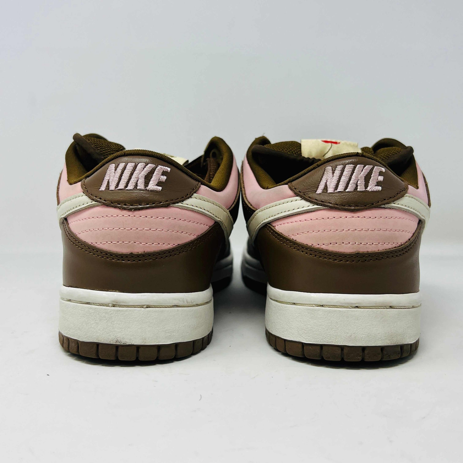 A 2005 Nike SB Dunk Low Stussy Cherry in brown and pink rests on its box, featuring a cream swoosh. The authentic shoe includes extra pink laces in a clear NIKE SB bag. The box label shows size 9.