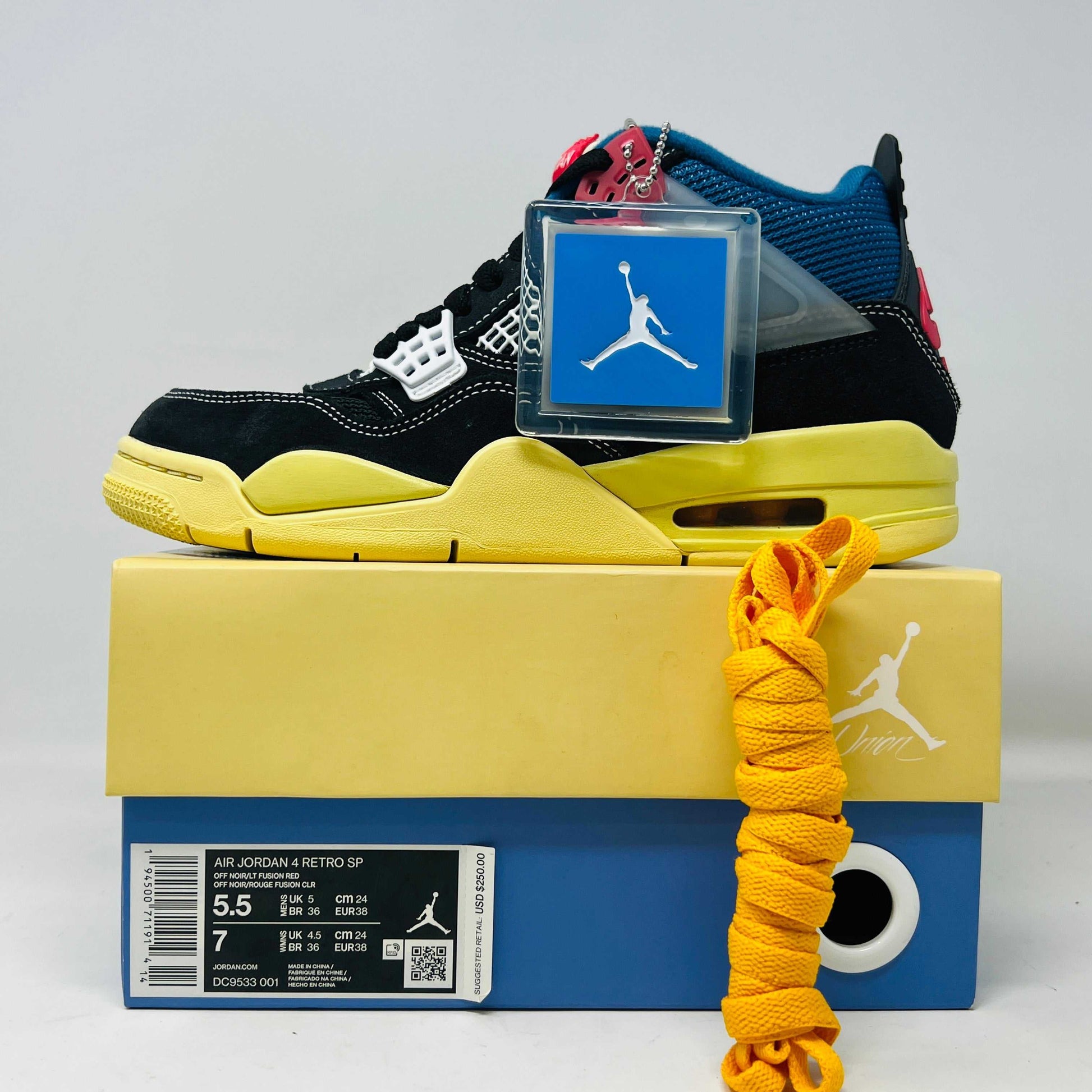 Displayed on their box, the 100% authentic Jordan 4 Retro Union Off Noir sneakers from the Jordan brand showcase clean black uppers, a yellow sole, blue accents, and feature a clear hang tag with a white Jumpman logo. Yellow extra laces are also placed on the box.