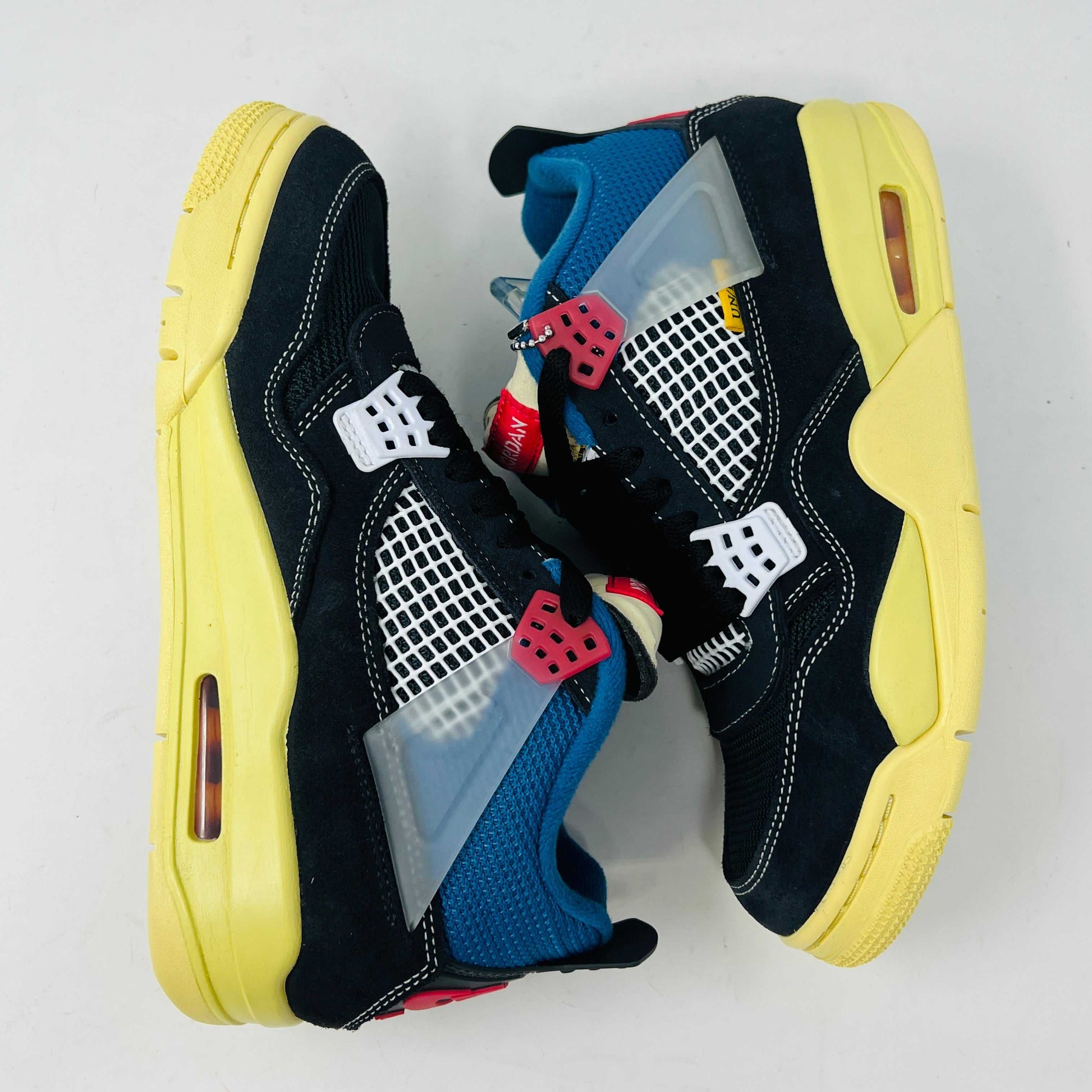 Displayed on their box, the 100% authentic Jordan 4 Retro Union Off Noir sneakers from the Jordan brand showcase clean black uppers, a yellow sole, blue accents, and feature a clear hang tag with a white Jumpman logo. Yellow extra laces are also placed on the box.