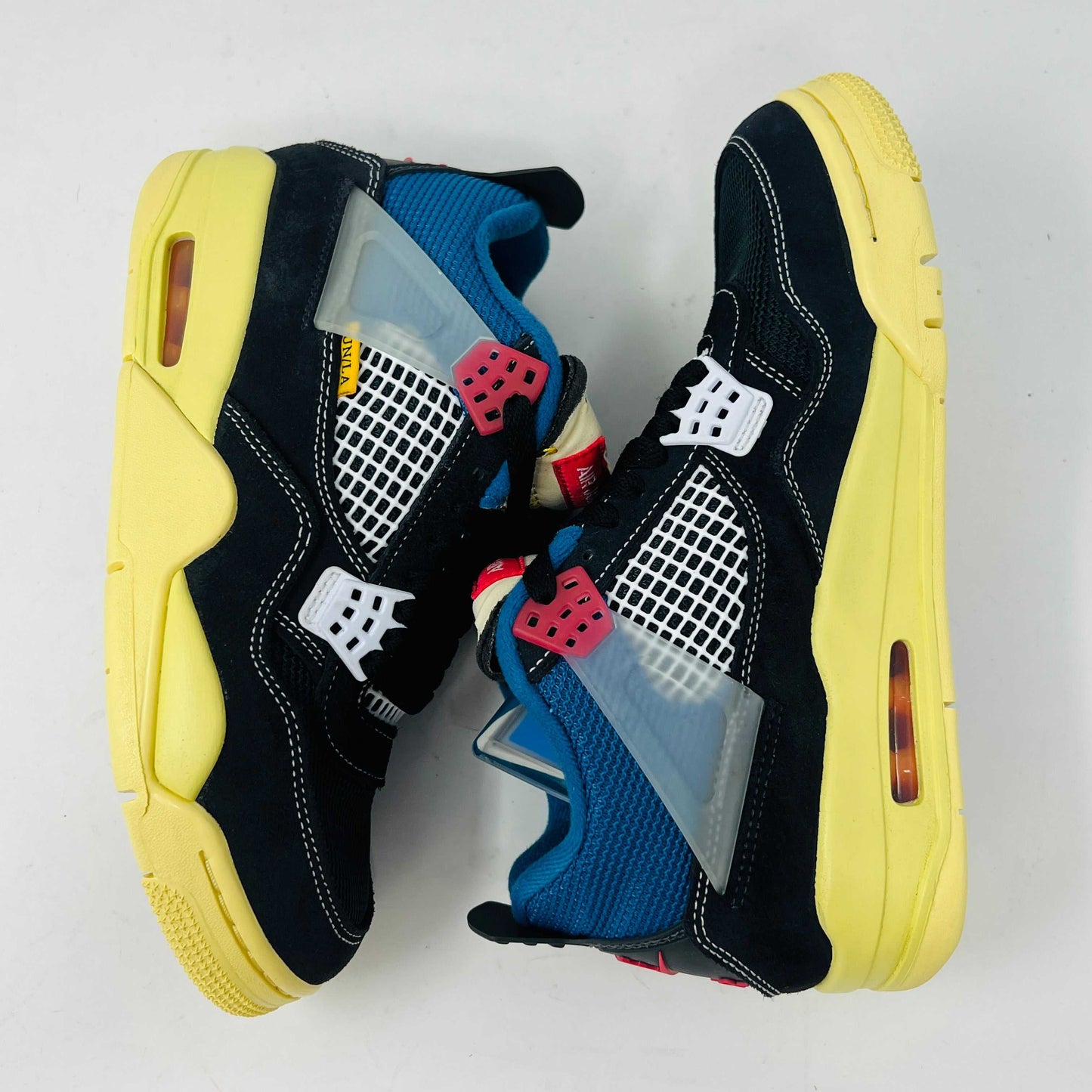 Displayed on their box, the 100% authentic Jordan 4 Retro Union Off Noir sneakers from the Jordan brand showcase clean black uppers, a yellow sole, blue accents, and feature a clear hang tag with a white Jumpman logo. Yellow extra laces are also placed on the box.