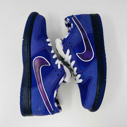 The Nike SB Dunk Low Concepts Purple Lobster sneaker, featuring a clean upper with a white swoosh, rests on a blue and white Nike SB shoebox. An extra bag with pink laces adds a vibrant touch in front of the box.