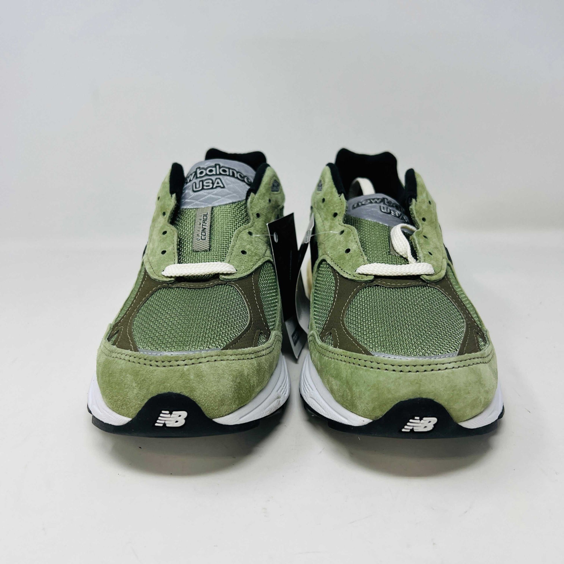 A New Balance 990v3 MiUSA JJJJound Olive sneaker from the 2022 collection is displayed on a white box featuring the brands logo. The olive shoe features a black N logo, white sole, grey accents, and extra green laces placed in front of the box.