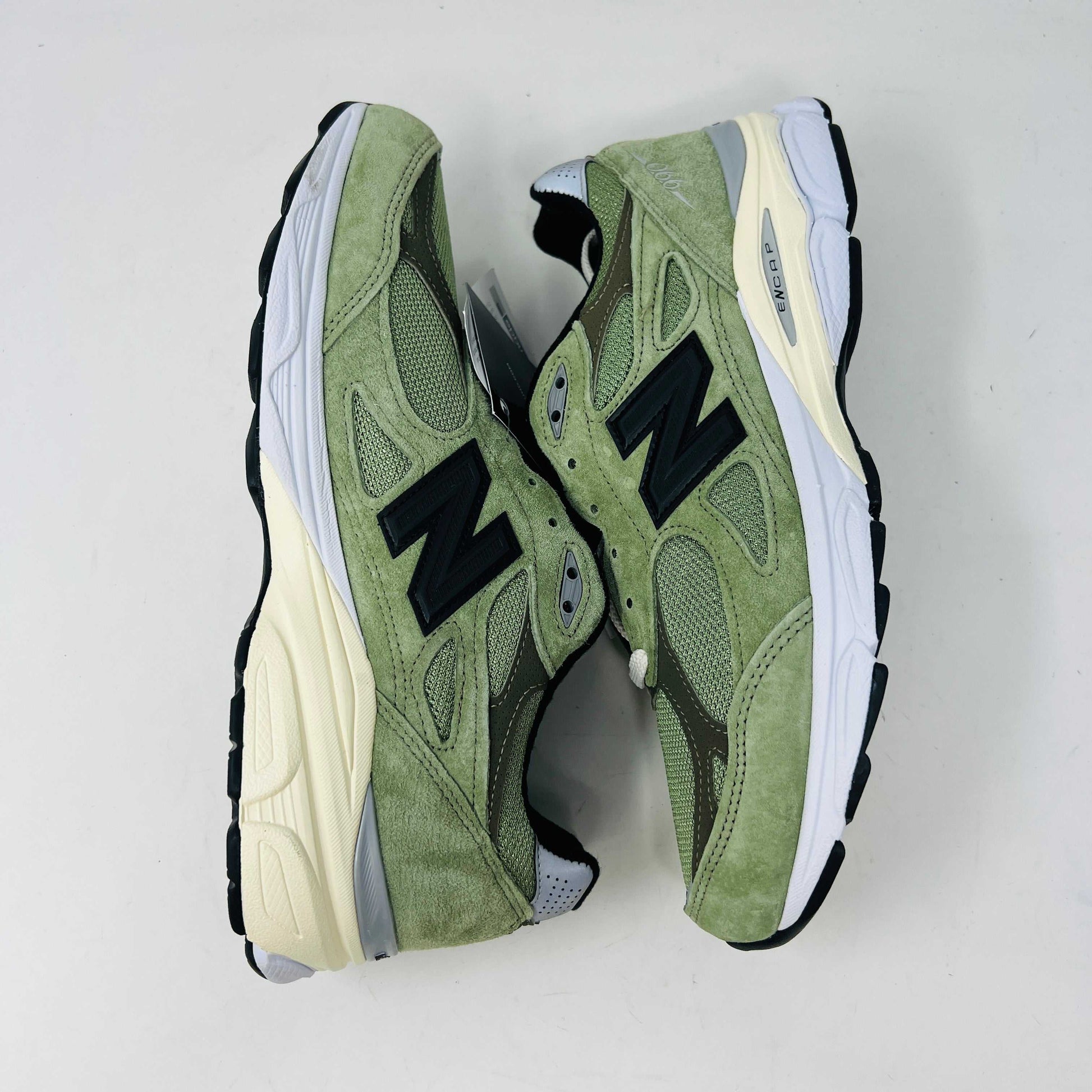 A New Balance 990v3 MiUSA JJJJound Olive sneaker from the 2022 collection is displayed on a white box featuring the brands logo. The olive shoe features a black N logo, white sole, grey accents, and extra green laces placed in front of the box.