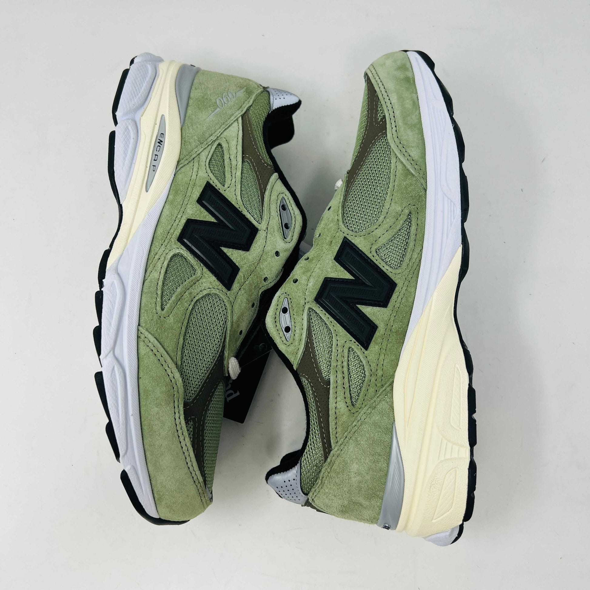 A New Balance 990v3 MiUSA JJJJound Olive sneaker from the 2022 collection is displayed on a white box featuring the brands logo. The olive shoe features a black N logo, white sole, grey accents, and extra green laces placed in front of the box.