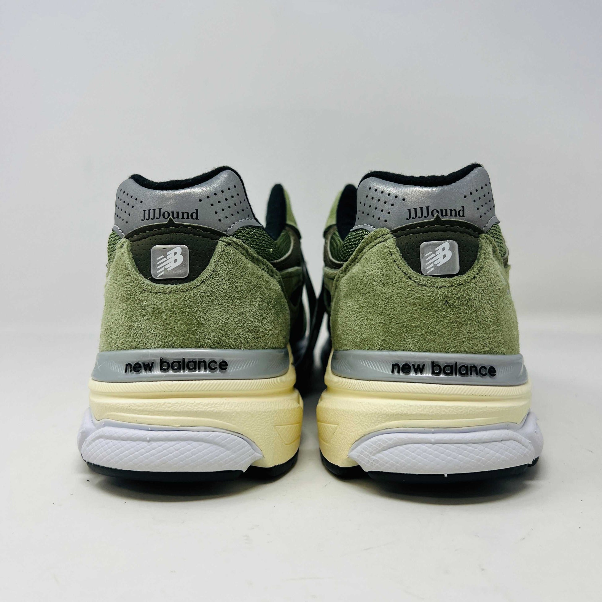 A New Balance 990v3 MiUSA JJJJound Olive sneaker from the 2022 collection is displayed on a white box featuring the brands logo. The olive shoe features a black N logo, white sole, grey accents, and extra green laces placed in front of the box.