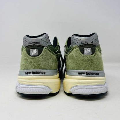 A New Balance 990v3 MiUSA JJJJound Olive sneaker from the 2022 collection is displayed on a white box featuring the brands logo. The olive shoe features a black N logo, white sole, grey accents, and extra green laces placed in front of the box.