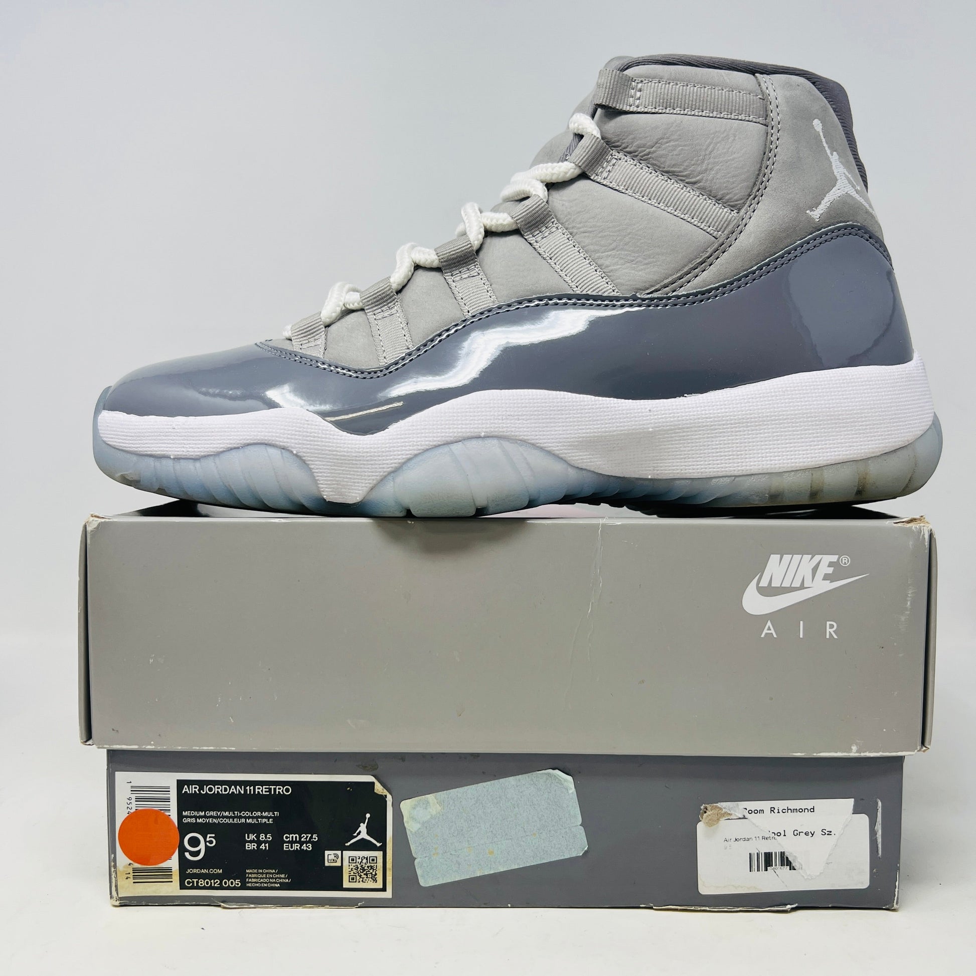 The Jordan 11 Retro Cool Grey (2021) sneaker, featuring clean gray and white uppers with glossy patent leather accents and white laces, is displayed on a gray Nike Air shoebox. The shoe is authentic in size 9.5, with the box in good condition.