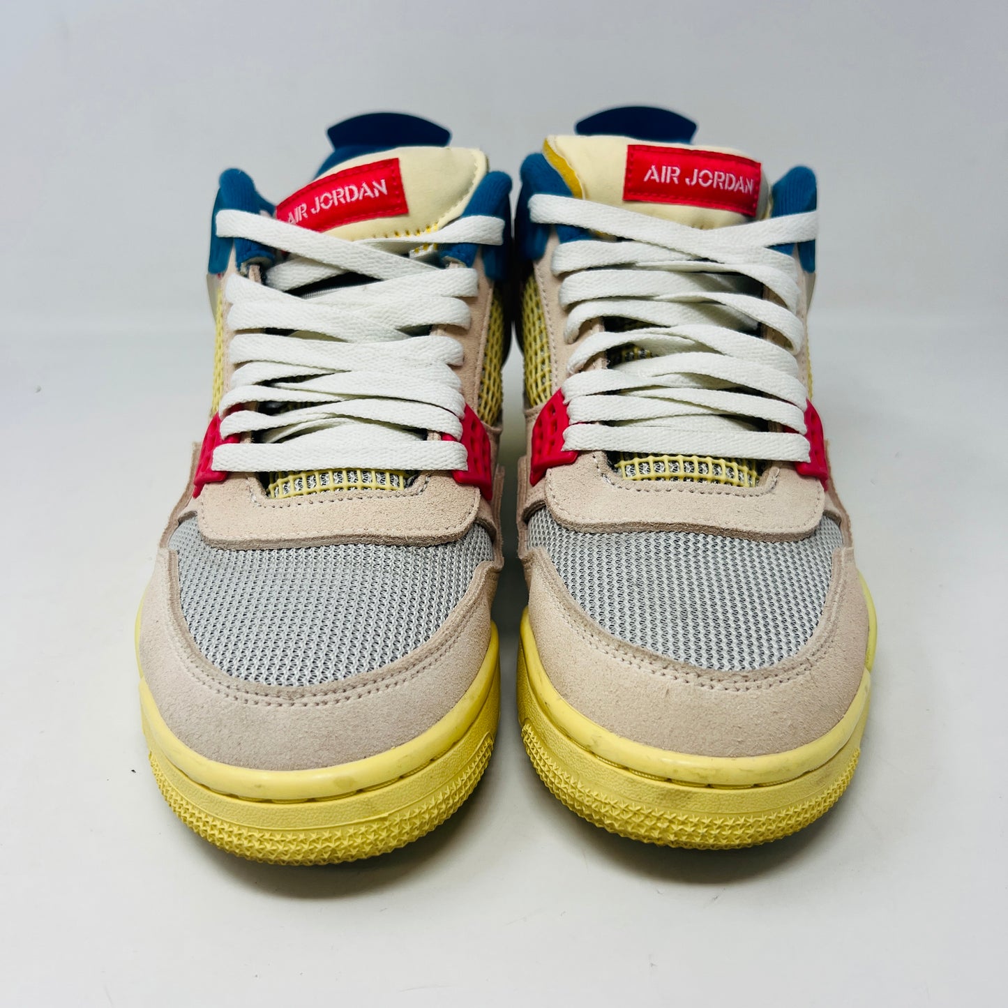 The Jordan 4 Retro Union Guava Ice sneaker from Jordan features a vibrant mix of beige, yellow, blue, and red on its clean uppers. It includes extra yellow laces packaged in a plastic bag alongside the shoe box.