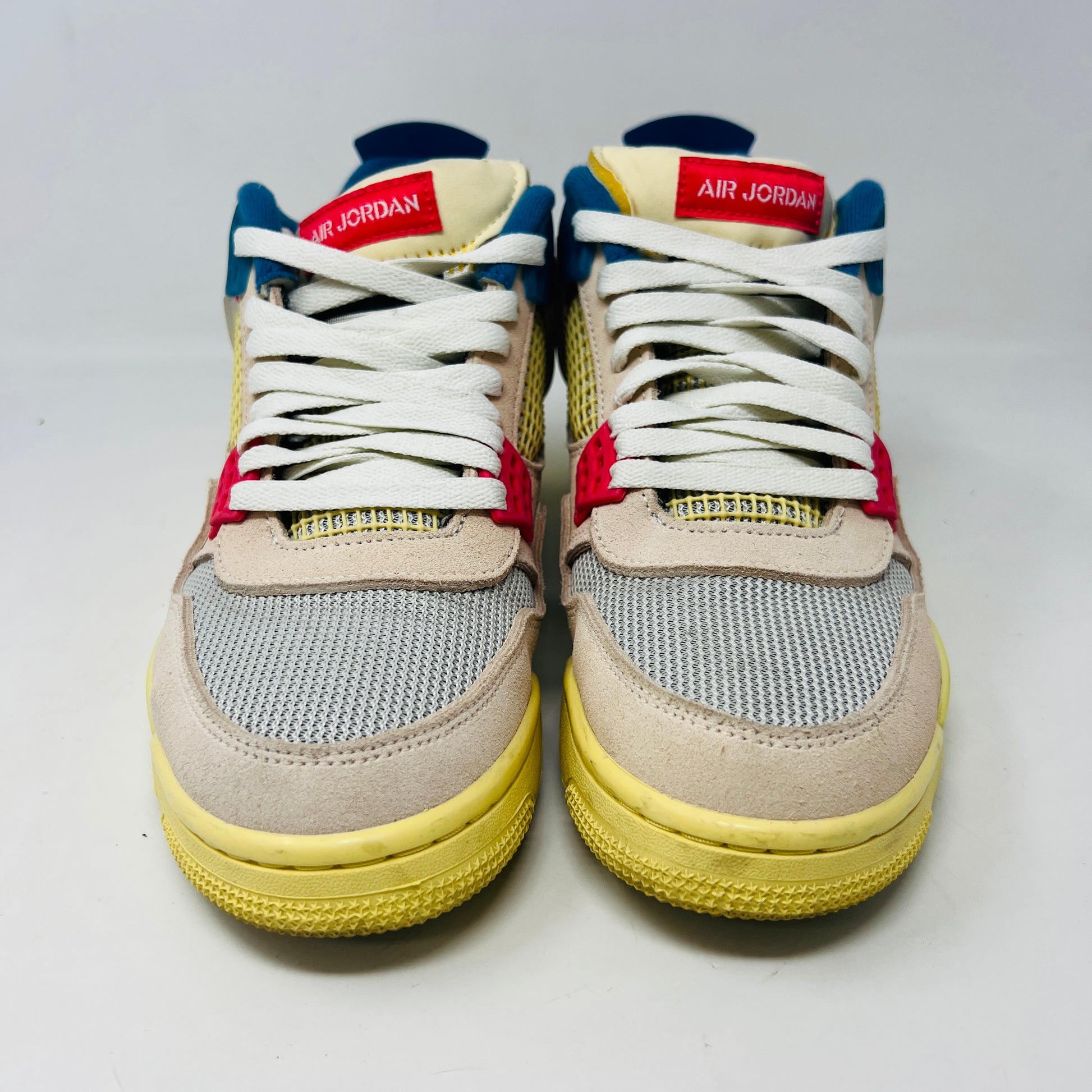 The Jordan 4 Retro Union Guava Ice sneaker from Jordan features a vibrant mix of beige, yellow, blue, and red on its clean uppers. It includes extra yellow laces packaged in a plastic bag alongside the shoe box.