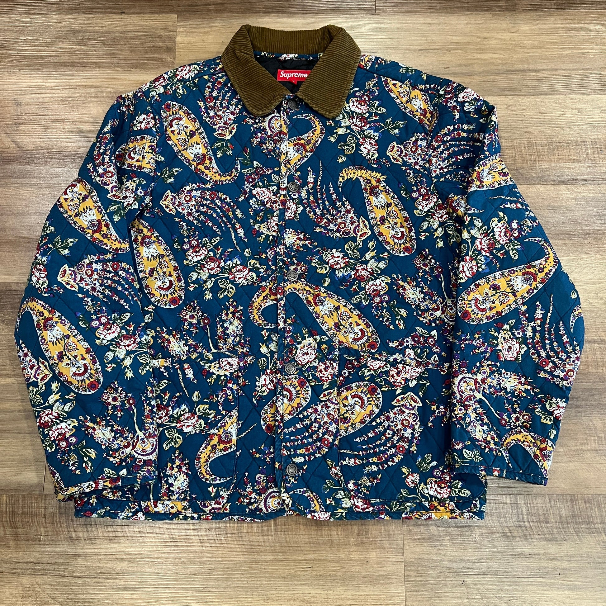 The preowned Supreme Quilted Paisley Jacket in Navy Paisley, featuring a medium floral paisley pattern and dark collar, lies flat on a wooden floor. Its ornate design showcases vibrant red, yellow, and white colors against a dark background.