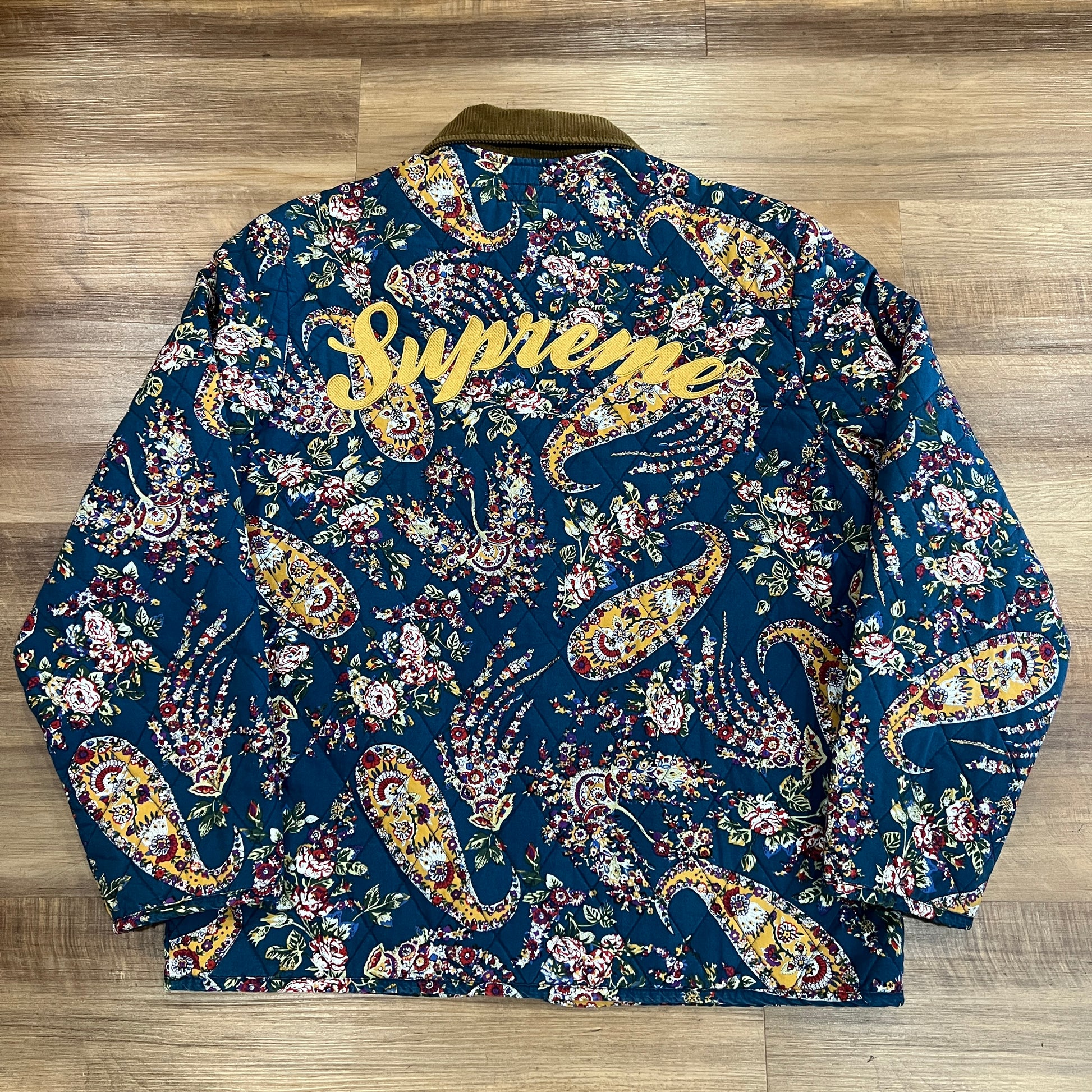 The preowned Supreme Quilted Paisley Jacket in Navy Paisley, featuring a medium floral paisley pattern and dark collar, lies flat on a wooden floor. Its ornate design showcases vibrant red, yellow, and white colors against a dark background.