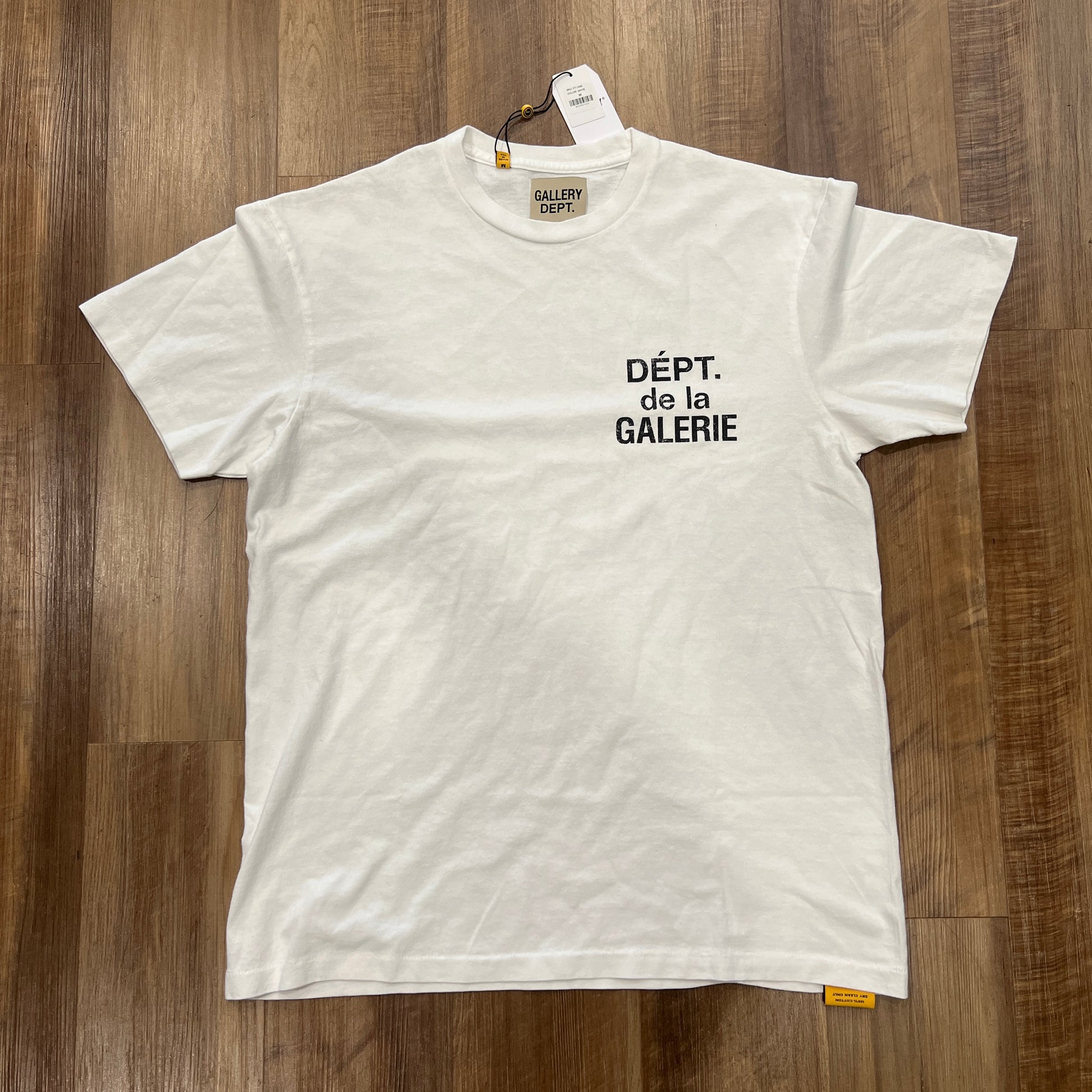 A new Gallery Dept. French Tee White is on a wooden floor, featuring black DÉPT. de la GALERIE text on the front and a tag near the collar.