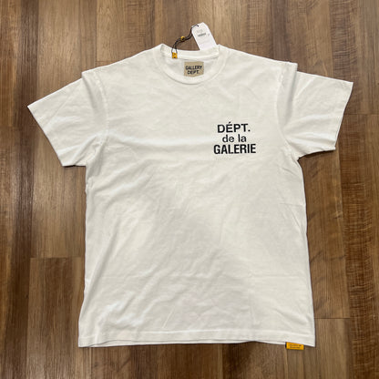 A new Gallery Dept. French Tee White is on a wooden floor, featuring black DÉPT. de la GALERIE text on the front and a tag near the collar.