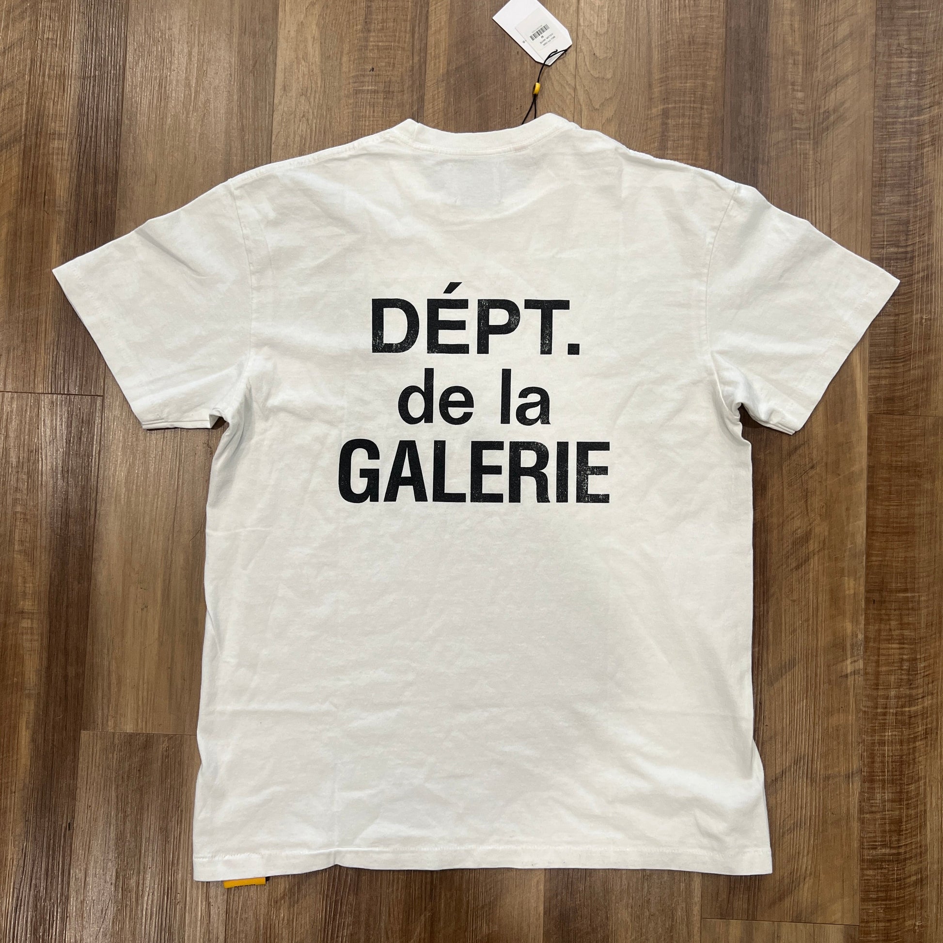A pristine Gallery Dept. French Tee White by Gallery Dept, displayed on a wooden floor, features DÉPT. de la GALERIE in bold black letters on the back with a tag attached to the collar.