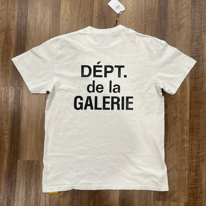 A pristine Gallery Dept. French Tee White by Gallery Dept, displayed on a wooden floor, features DÉPT. de la GALERIE in bold black letters on the back with a tag attached to the collar.