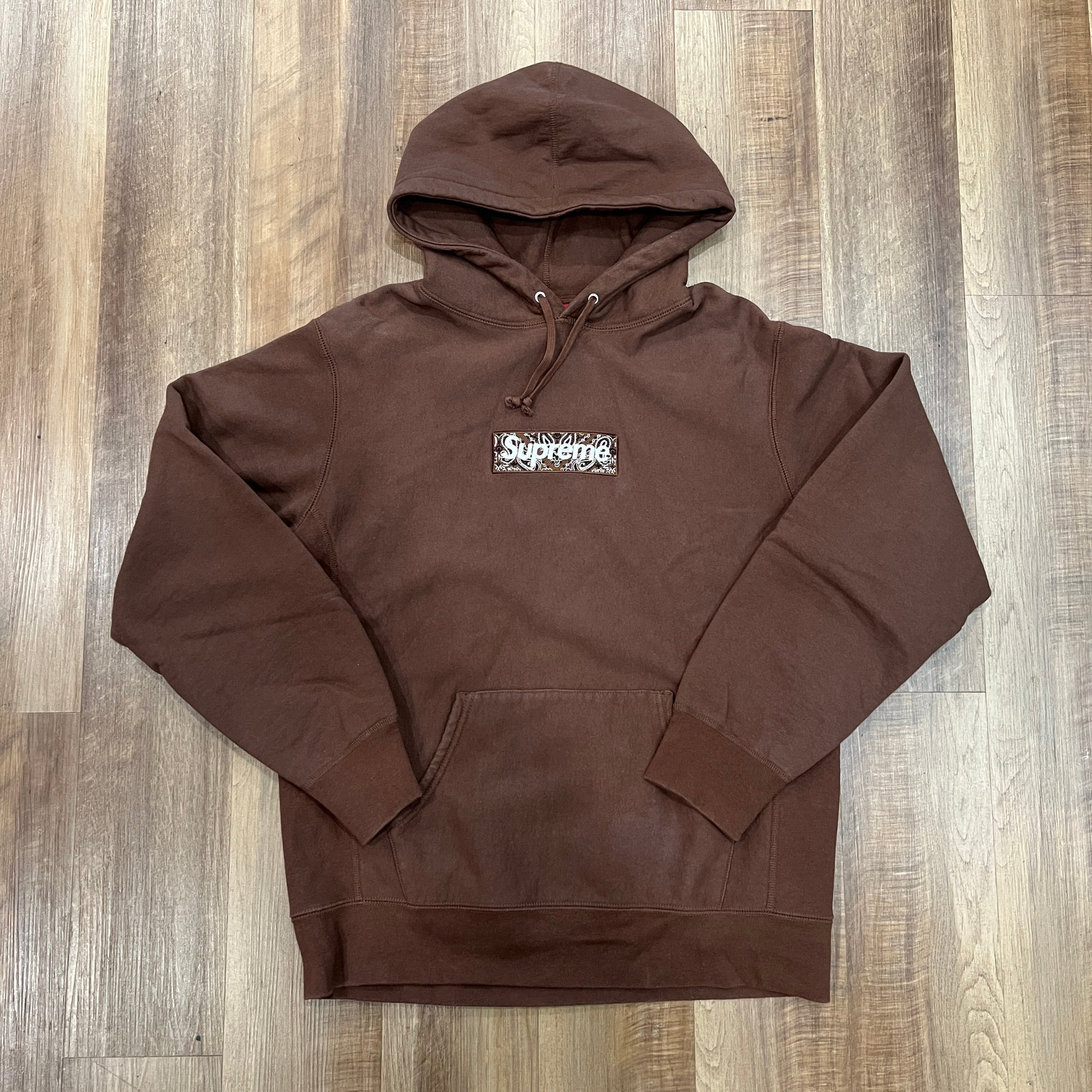 The Supreme Bandana Box Logo Hooded Sweatshirt in Dark Brown is preowned, featuring a kangaroo pocket, drawstrings, and a chest logo patch. Displayed on an earthy-toned wooden floor, it exudes timeless appeal.