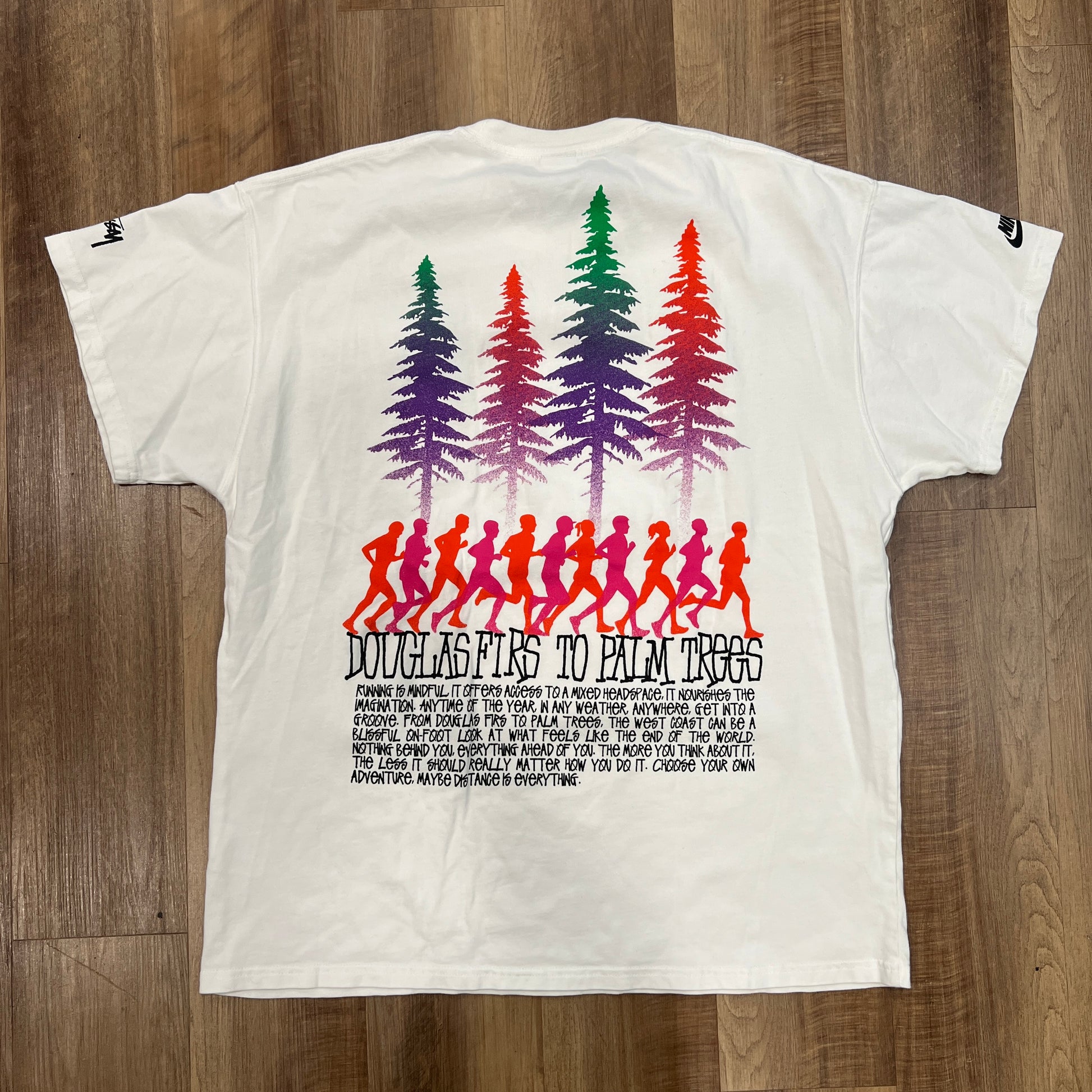 A new white Nike x Stussy Douglas Firs to Palm Trees T-shirt, by Stussy, showcasing a vibrant design of colorful palm trees above red silhouettes of people running, laid flat on a wooden floor.