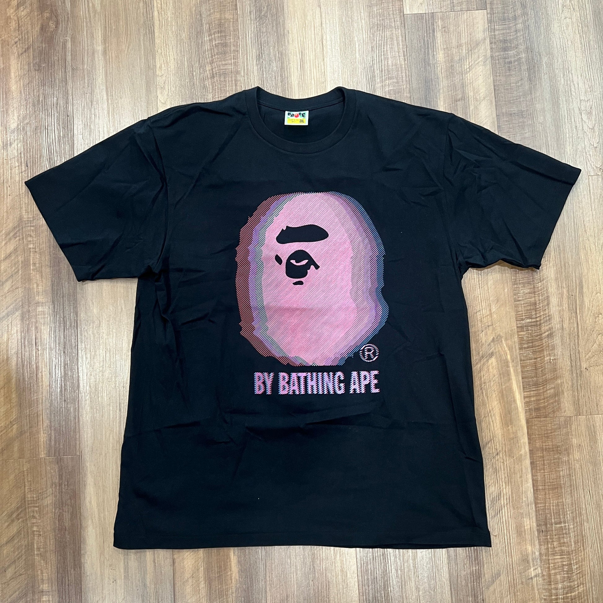 The BAPE Glitch Art Tee Black by Bape is showcased on a wooden floor, featuring a stylized pink ape face graphic with BY BATHING APE elegantly printed below.
