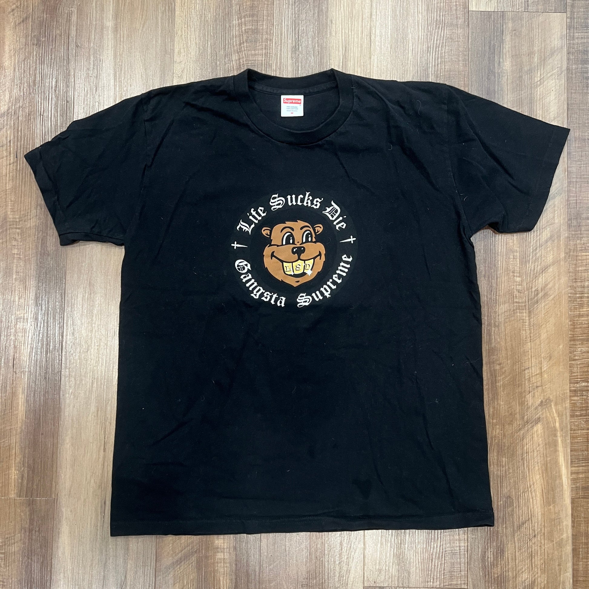 Preowned Supreme Life Sucks Die Tee Black on wooden floor featuring a cartoon bear face logo with Life Sucks Die above, Gangsta Supreme below, and the bear wearing a gold chain.
