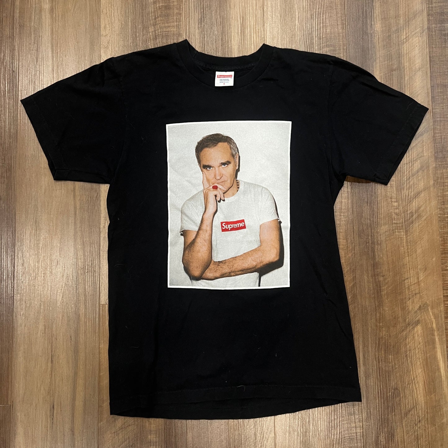 This preowned Supreme Morrissey Tee Black features an image of a person in a white shirt with the iconic red and white Supreme logo, exuding casual style with a touch of streetwear edge, perfectly laid flat on a wooden floor.